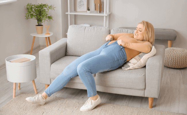 a photo of a woman lying down in the sofa trying to close the zipper of her very tight jeans