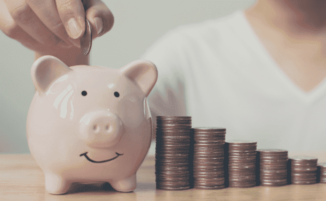 a photo of a piggy bank with a pile of coins on the side and a hand dropping a coin on the piggy bank.