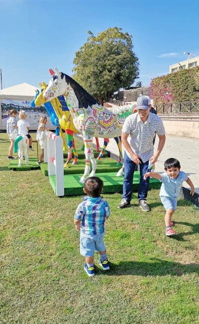 father having fun with his two toddler boys with colorful statues of horses at the back