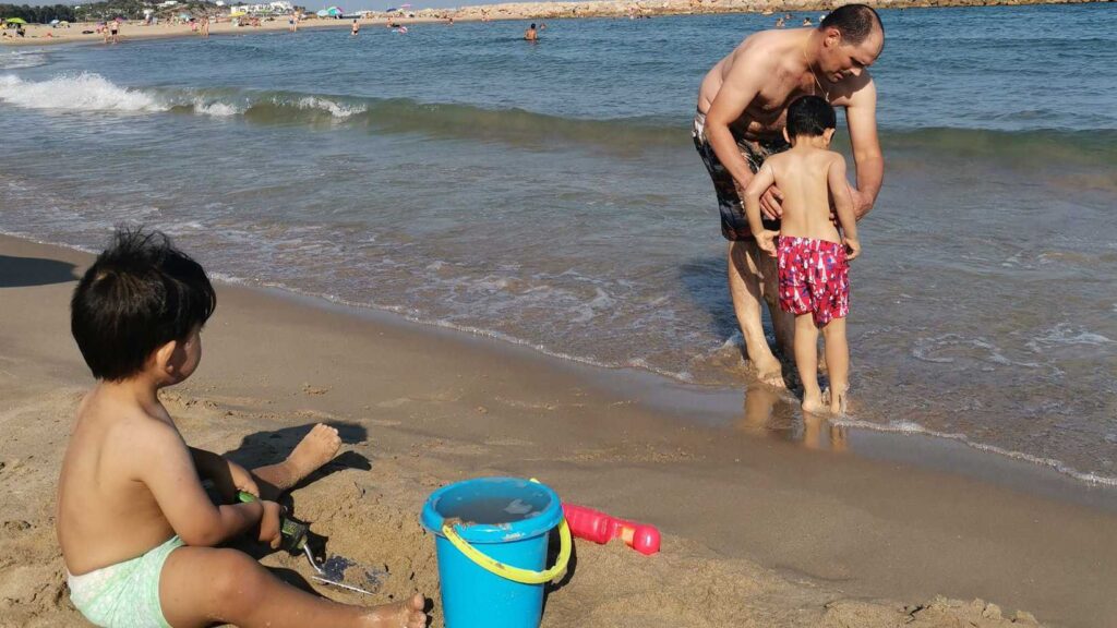 a photo of a kid playing in the sand and a father helping his other son dip into the water for a swim.