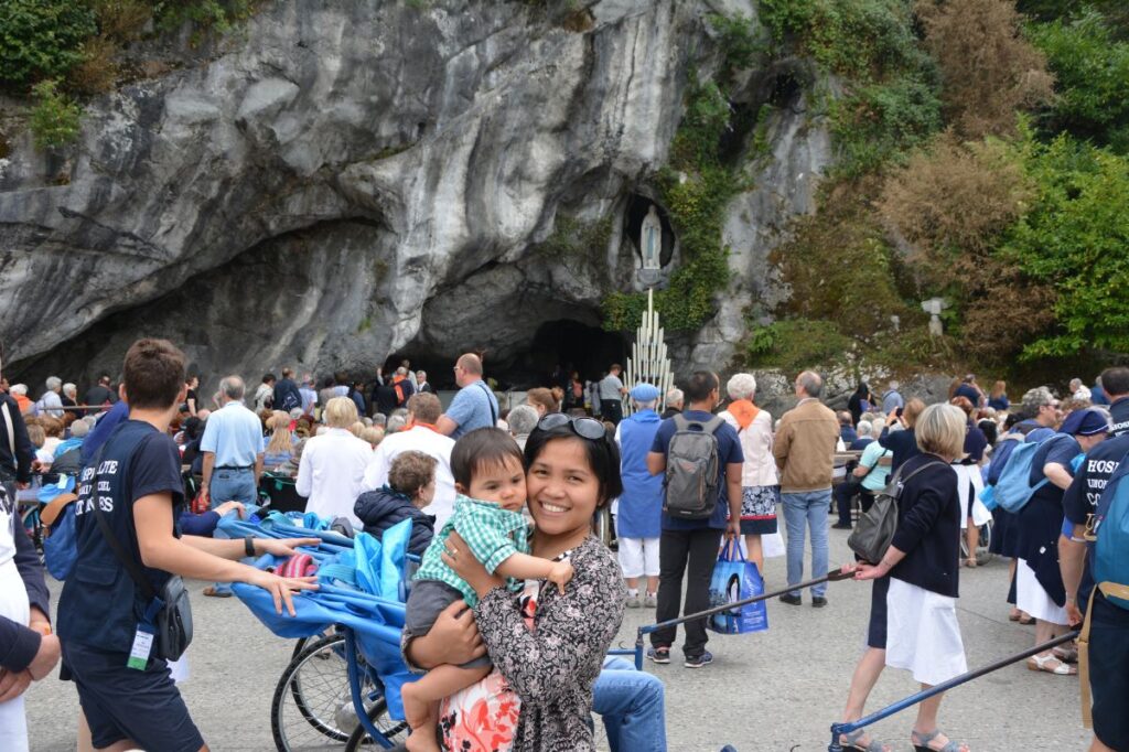 Photo of a woman holding a child in front of the Grotto of Lourdes in Lourdes, France, a well-known Catholic pilgrimage site, with many other visitors gathered for prayer and reflection near the grotto.