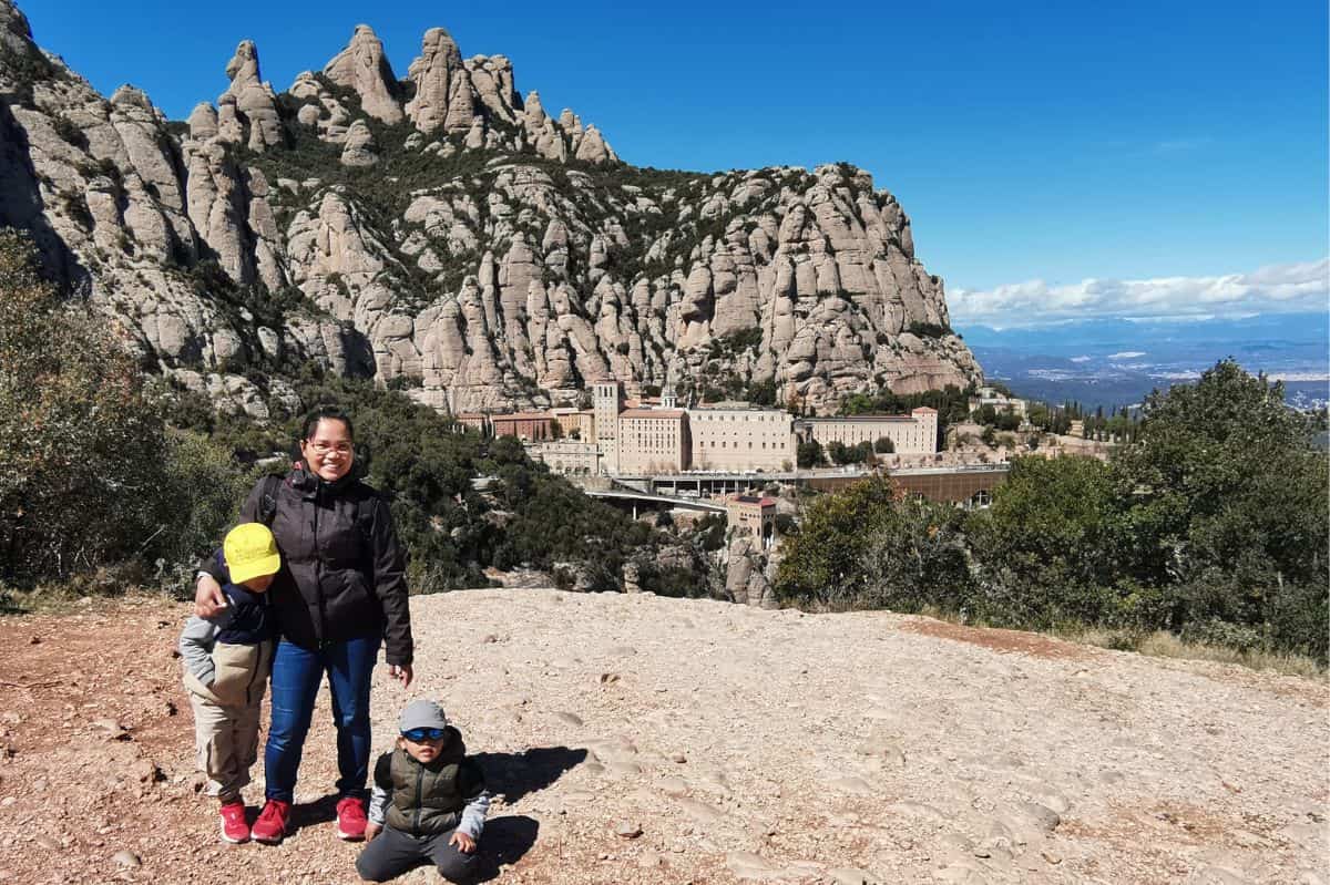 A family stands on a rocky overlook with the Montserrat Monastery and its dramatic mountainous backdrop in the distance. The clear blue sky and rugged terrain highlight the natural beauty surrounding this renowned Catholic pilgrimage site in Catalonia, Spain.
