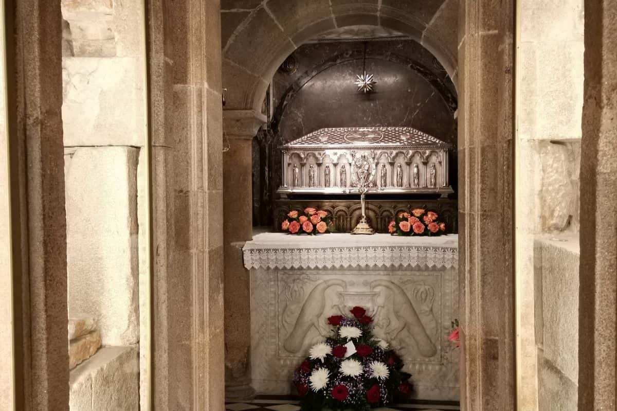 Interior view of the crypt in Santiago de Compostela Cathedral, housing the ornate silver reliquary of St. James the Apostle. The reliquary is adorned with intricate carvings and surrounded by fresh floral arrangements of roses and white blooms, creating a solemn and sacred atmosphere.