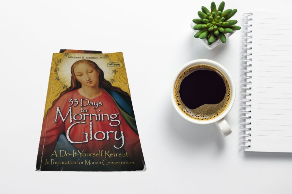 A flat-lay image of the book "33 Days to Morning Glory: A Do-It-Yourself Retreat In Preparation for Marian Consecration" by Michael E. Gaitley, MIC. The book, featuring an illustration of the Virgin Mary on the cover, is placed next to a cup of black coffee, a small green succulent plant, and a lined notebook on a white background.
