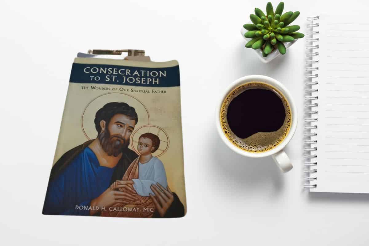 A flat-lay image featuring the book "Consecration to St. Joseph: The Wonders of Our Spiritual Father" by Donald H. Calloway, MIC. The book, which has a cover illustration of St. Joseph holding baby Jesus, is placed next to a cup of black coffee, a small green succulent plant, and a lined notebook on a white background.