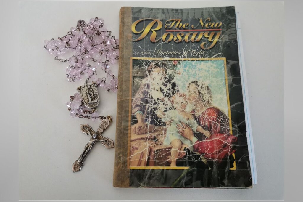 A well-worn copy of "The New Rosary: Mysteries of Light" placed on a flat surface next to a set of pink crystal rosary beads. The rosary features a silver crucifix and a medallion with a religious depiction. The book's cover shows a faded image of the Holy Family, giving it a vintage and used appearance.