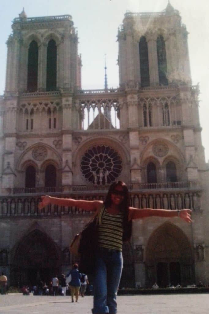 A woman stands with arms outstretched in front of the iconic twin towers of Notre-Dame Cathedral in Paris, one of the most renowned Catholic pilgrimage sites in France. The historic Gothic architecture and detailed rose window loom grandly behind her under a clear sky.
