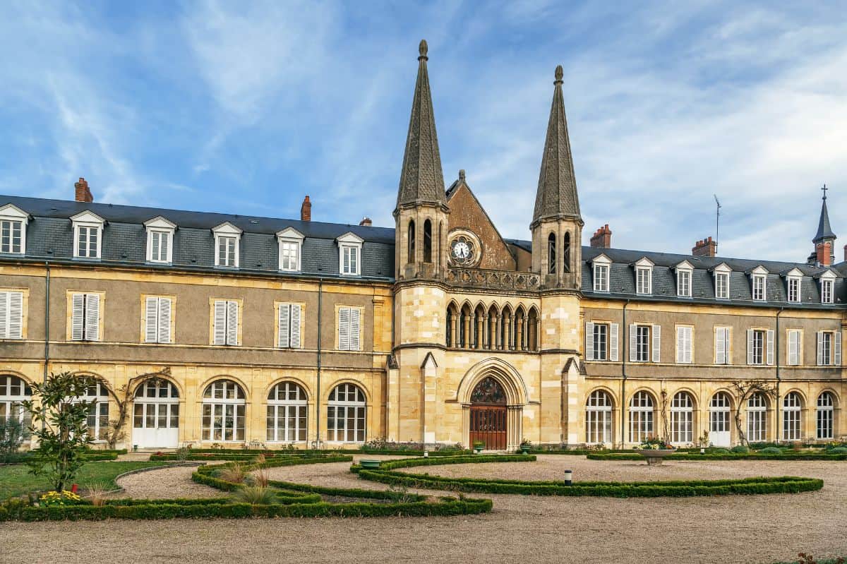 A symmetrical view of the St. Gildard Convent in Nevers, France, featuring twin spires, a central rose window, and rows of shuttered windows above graceful archways. The neatly trimmed garden paths and sunny sky add a peaceful atmosphere to the historic site.