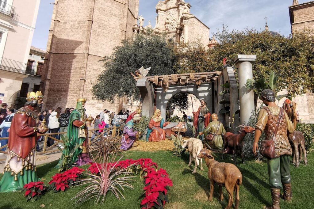 A vibrant outdoor Nativity scene set (signify the coming of Christ - one of the traditional Catholic monthly devotions for the month of December) against a historic stone church building. The display features life-sized figures of Mary, Joseph, baby Jesus in the manger, the Three Wise Men bearing gifts, shepherds, and animals like sheep and goats, surrounded by lush greenery and red poinsettias. Crowds of people are gathered in the background, enjoying the festive display on a sunny day.