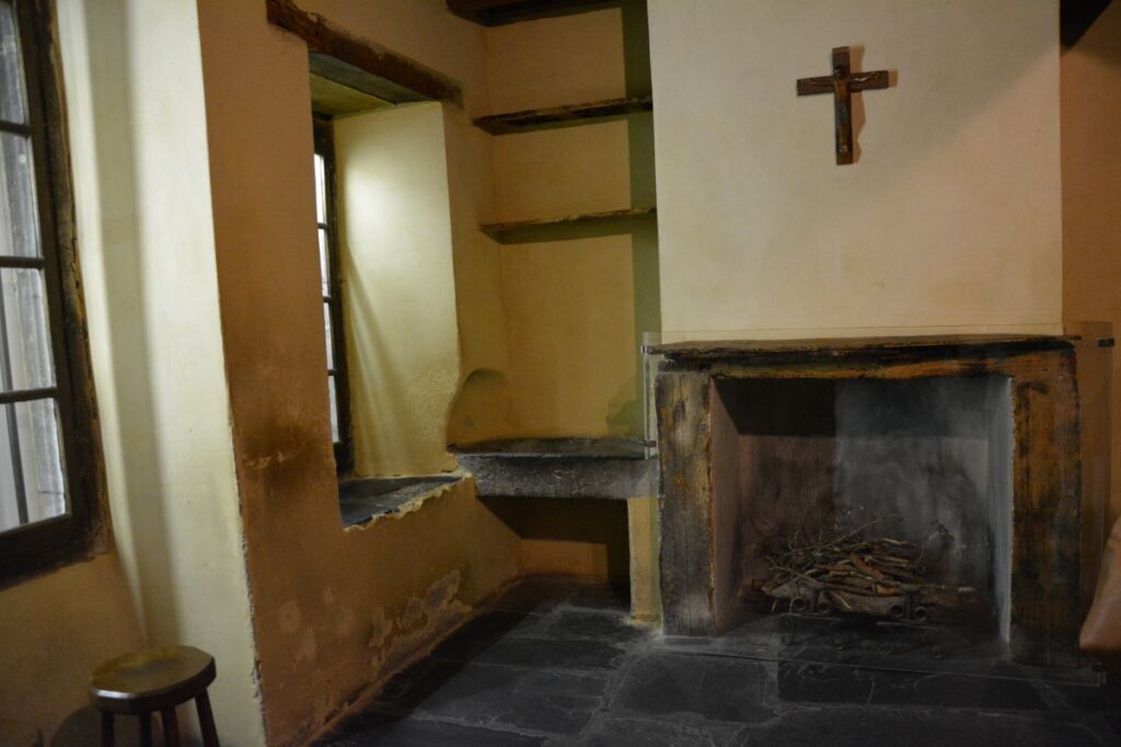 Interior view of Le Cachot - a significant place that is part of Our Lady of Lourdes Apparition Story to St. Bernadette Soubirous, the small, simple room where St. Bernadette and her family lived in Lourdes. The space features a rustic fireplace with a small pile of wood, a cross hanging above it, and a worn stone sink near a window, reflecting the family's humble living conditions.