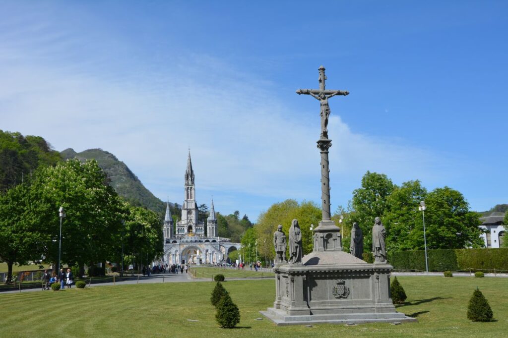 A wide view of the Sanctuary of Our Lady of Lourdes in France, featuring a crucifix and statues in the foreground with the basilica's spire rising against a clear blue sky. The scene is framed by lush green trees and rolling hills, evoking a peaceful and sacred atmosphere.