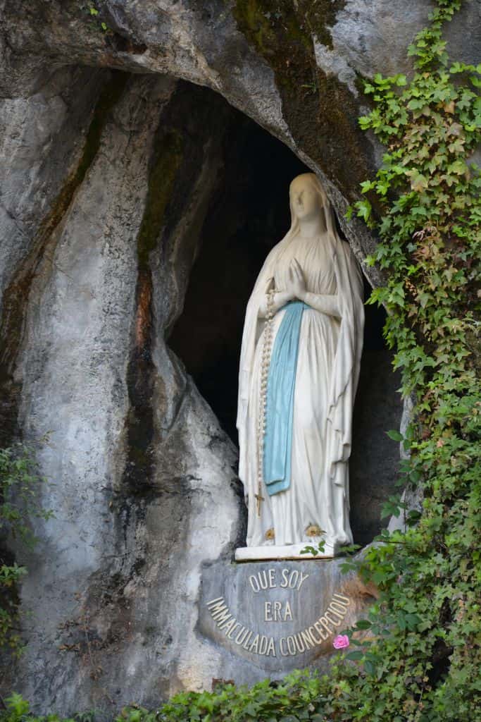 The statue of the Virgin Mary at the Grotto of Lourdes, depicted in white robes with a blue sash, holding a rosary. The inscription below reads 'Que soy era Immaculada Concepciou,' meaning 'I am the Immaculate Conception,' surrounded by moss-covered rocks and ivy, symbolizing the site of her apparition to St. Bernadette.
